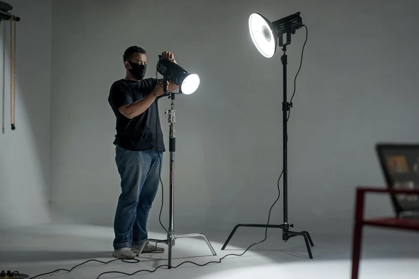 En person som bär ansiktsmask justerar en studiolampa i en fotostudio. Det finns ytterligare en upplyst studiolampa på ett stativ bredvid honom.