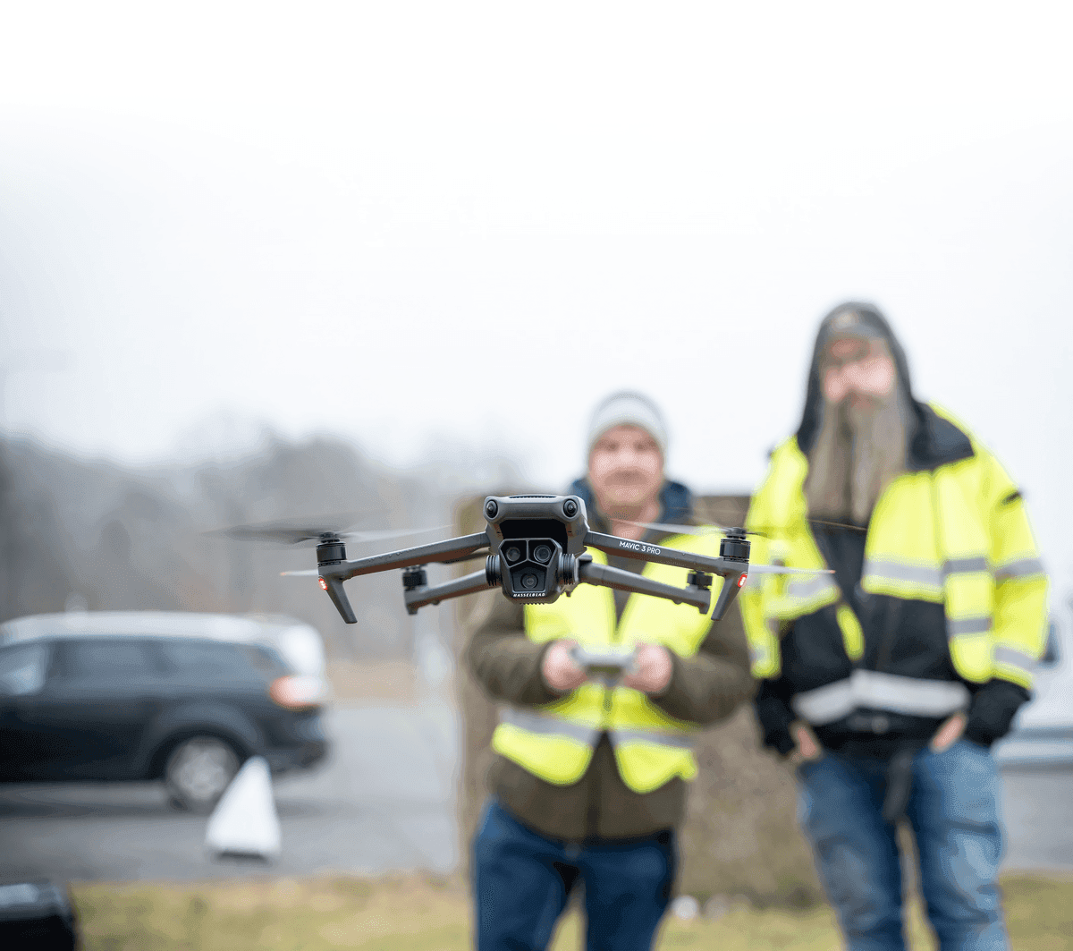 En drönare svävar i luften medan två personer i reflexvästar står i bakgrunden och kontrollerar den.