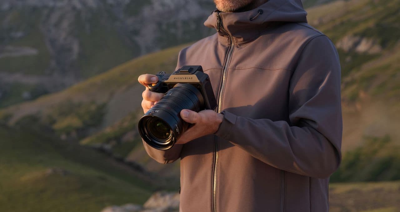 En person i en jacka håller en Hasselblad X2D II 100C-kamera med ett stort objektiv, med en bergig landskap i bakgrunden.