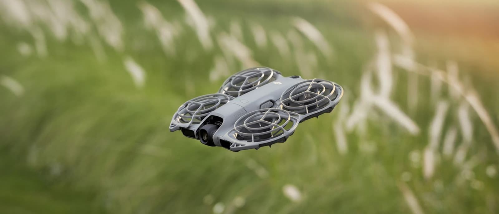 En DJI Neo 2-drönare svävar över ett grönt fält i dagsljus, omgiven av högt gräs och en suddig bakgrund.