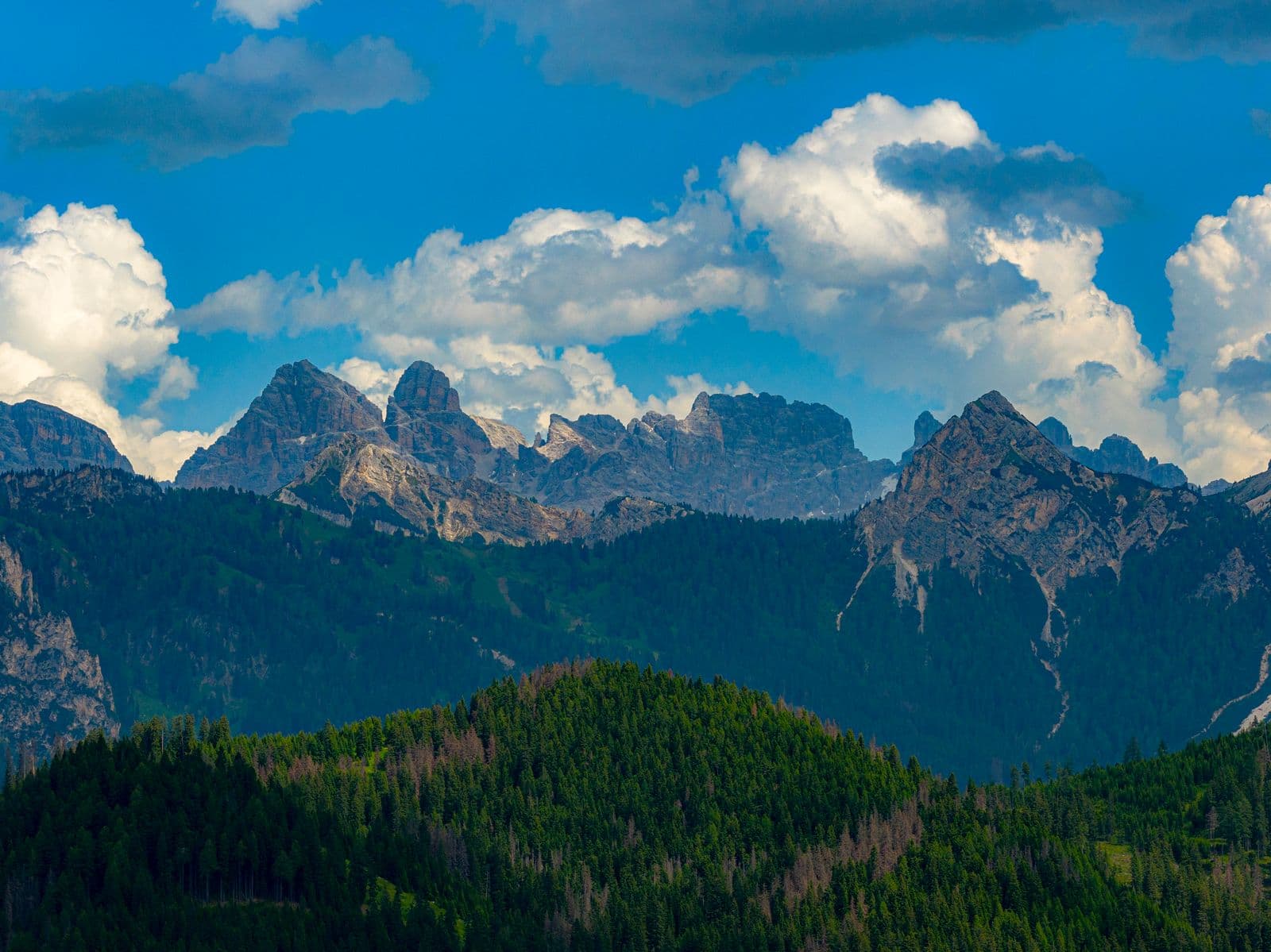 Drönarfoto taget med DJI Mavic 4 Pro: Majestic mountain peaks rise above lush green forests under a sky filled with dramatic clouds.