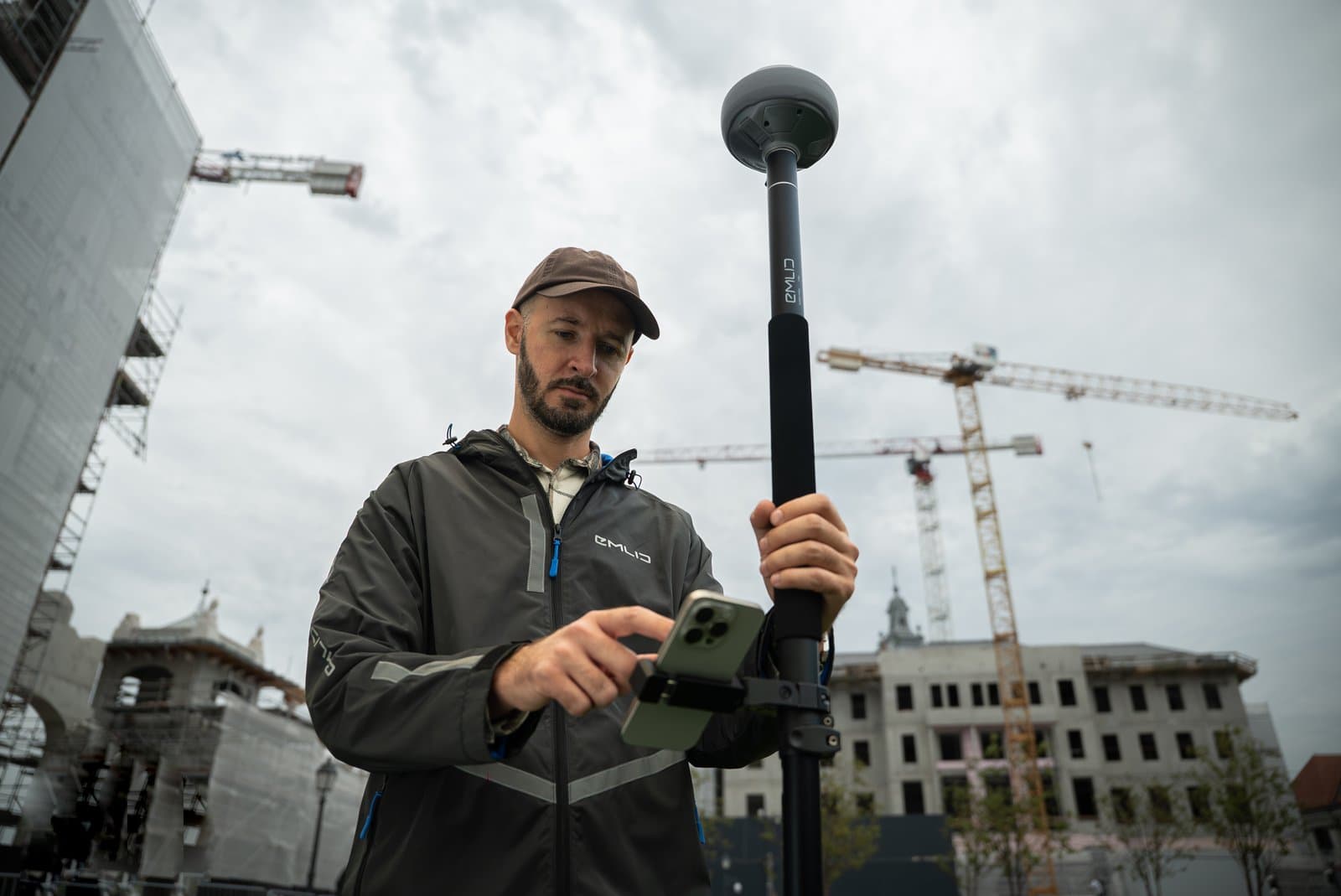 En man använder en GPS-enhet på en byggarbetsplats, omgiven av kranar och byggnader under konstruktion.