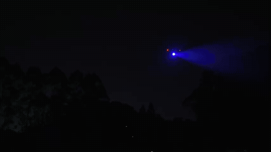 En drönare med starkt blått ljus svävar över ett mörkt skogslandskap på natten.