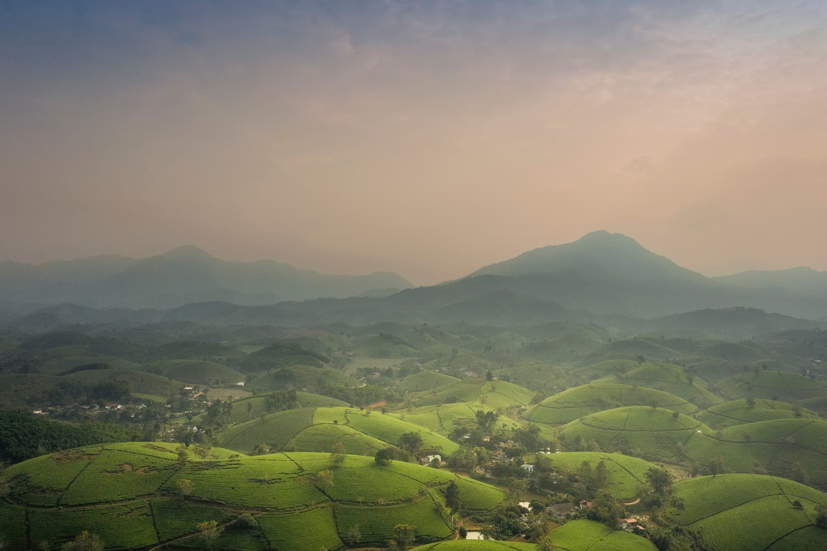 Gröna kullar sträcker sig över landskapet under en dimmig himmel, med berg som reser sig i bakgrunden.