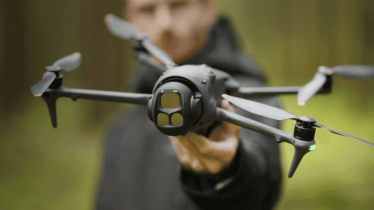 En person håller en avancerad drönare med flera rotorblad i en skogsmiljö.