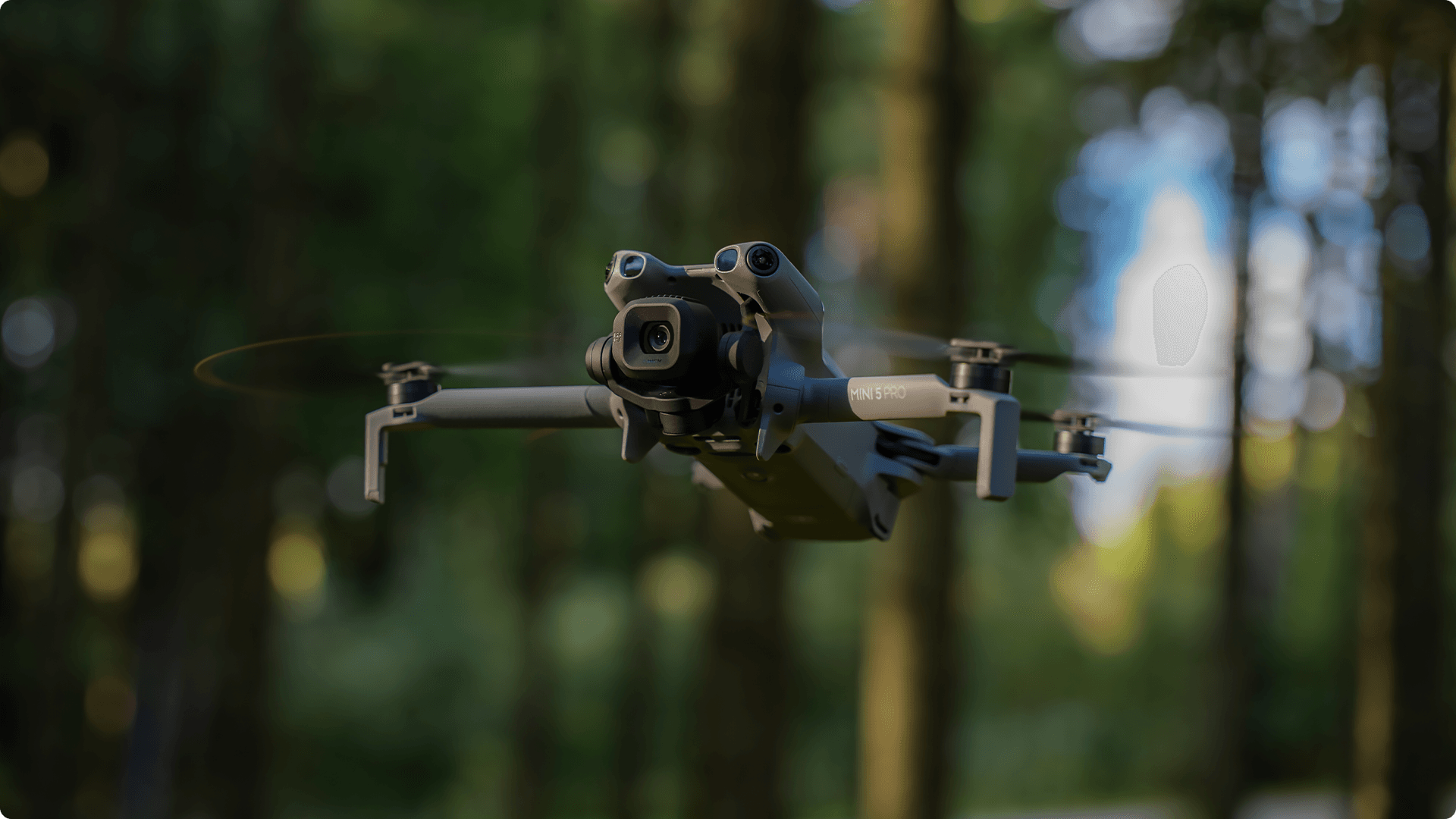 En DJI Mini 5 Pro-drönare svävar genom en skogsmiljö, med träd suddigt i bakgrunden.