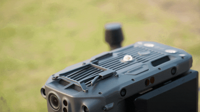 En drönare lyfter från marken och svävar uppåt, med en grönskande bakgrund i bakgrunden.