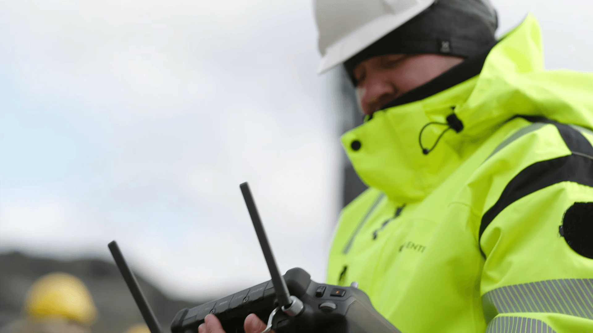 En person i skyddskläder och hjälm hanterar en fjärrkontroll utomhus, troligen för att styra en drönare.