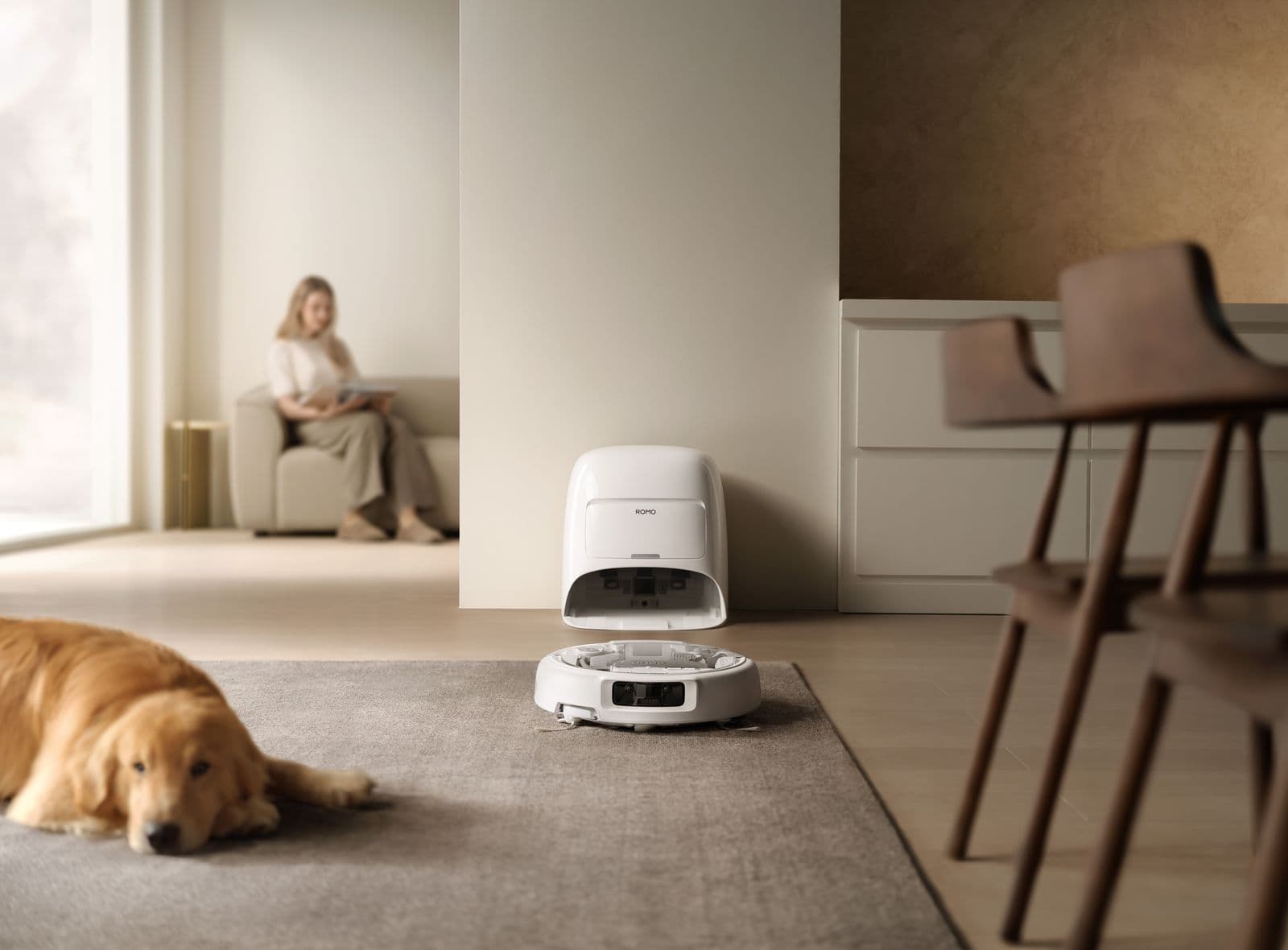 En robotdammsugare vid sin laddningsstation i ett modernt vardagsrum, med en hund som vilar på mattan och en person som sitter i bakgrunden.