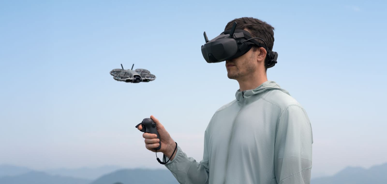 En person med VR-headset styr en DJI Neo 2-drönare med en handkontroll mot en bakgrund av berg och blå himmel.