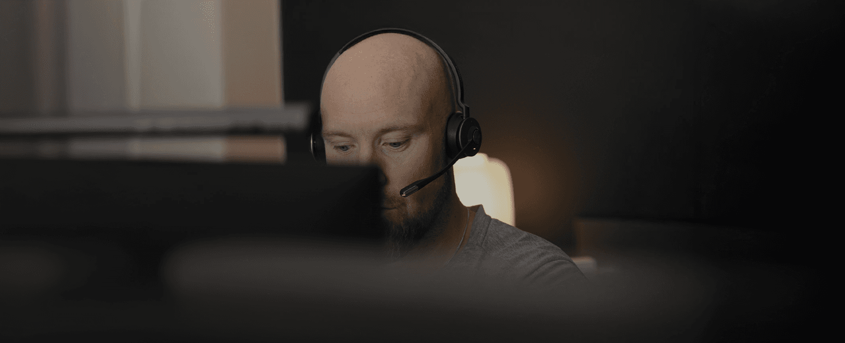 En person med headset arbetar fokuserat vid en dator i en mörk arbetsmiljö.