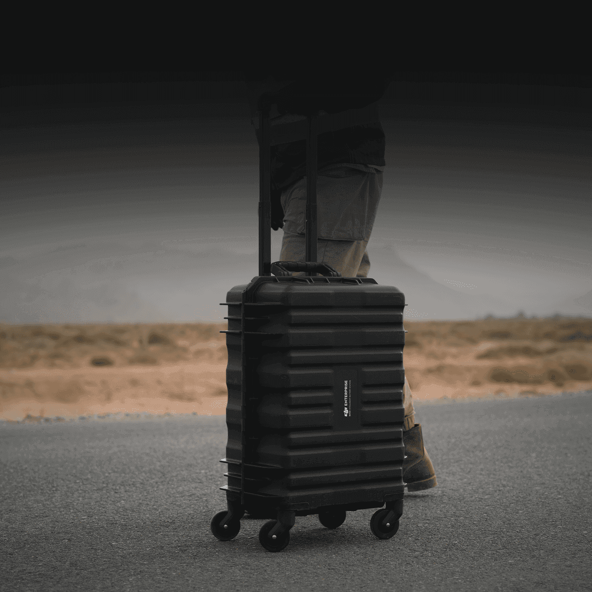 En person drar en robust svart resväska med hjul längs en öde väg i ett kargt landskap.