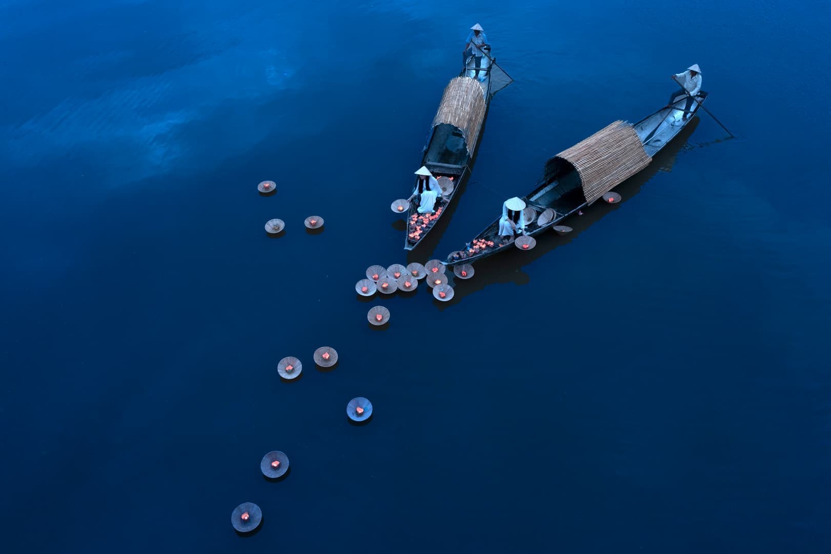 Två båtar med halmtak flyter på en lugn blå sjö, omgivna av flytande lyktor.