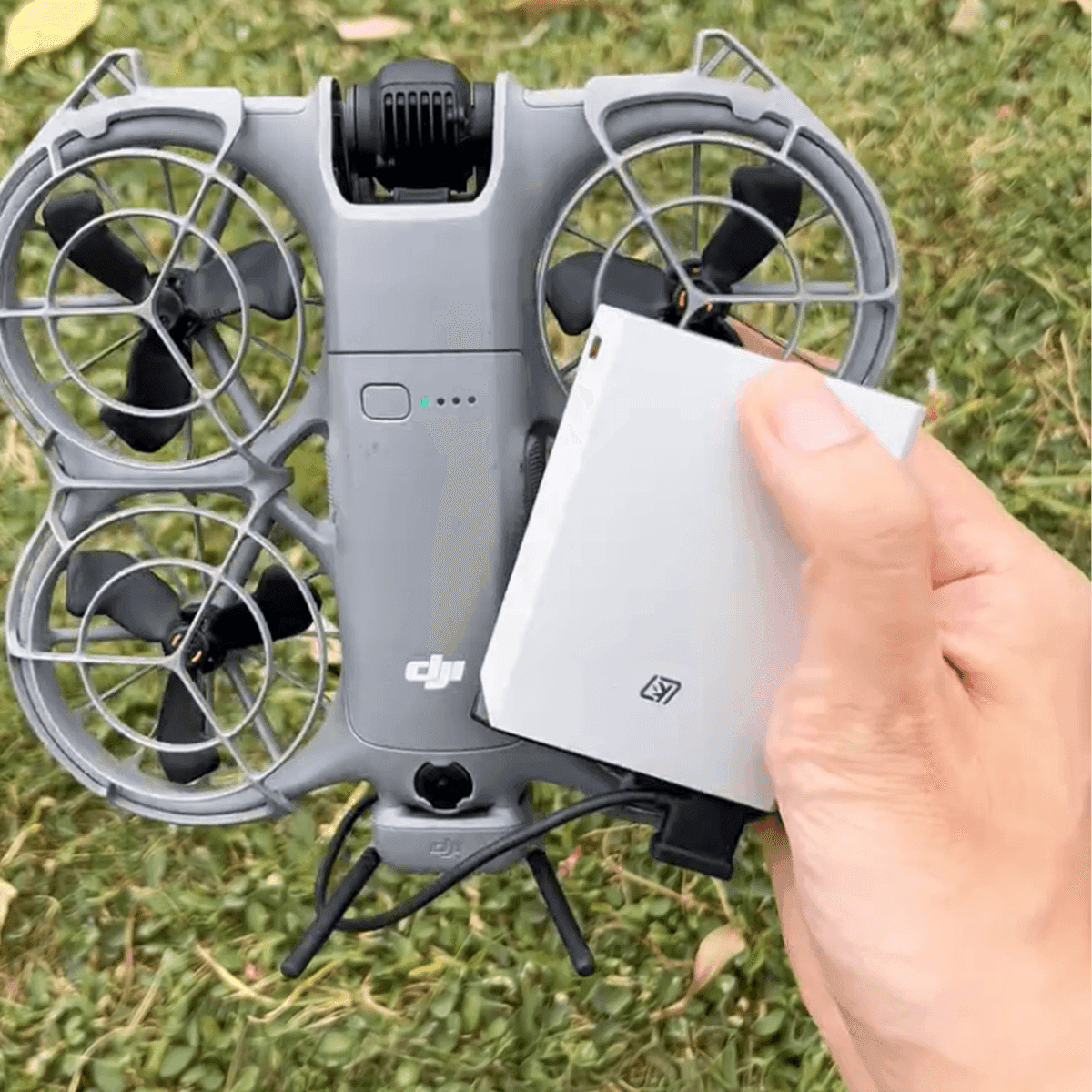 En hand håller i ett batteri bredvid en kompakt drönare med skyddade propellrar, placerad på gräs.