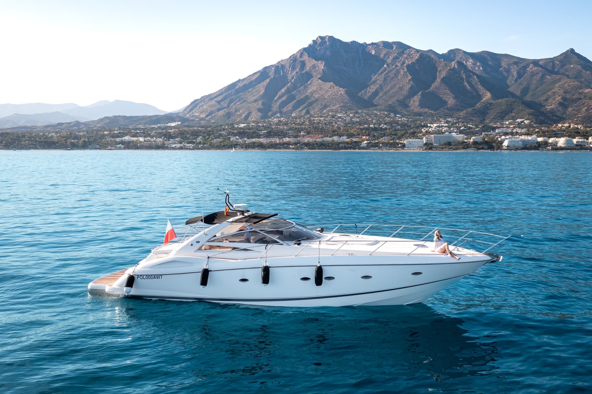 En lyxig vit yacht flyter på det klara blå vattnet med berg och en kuststad i bakgrunden.