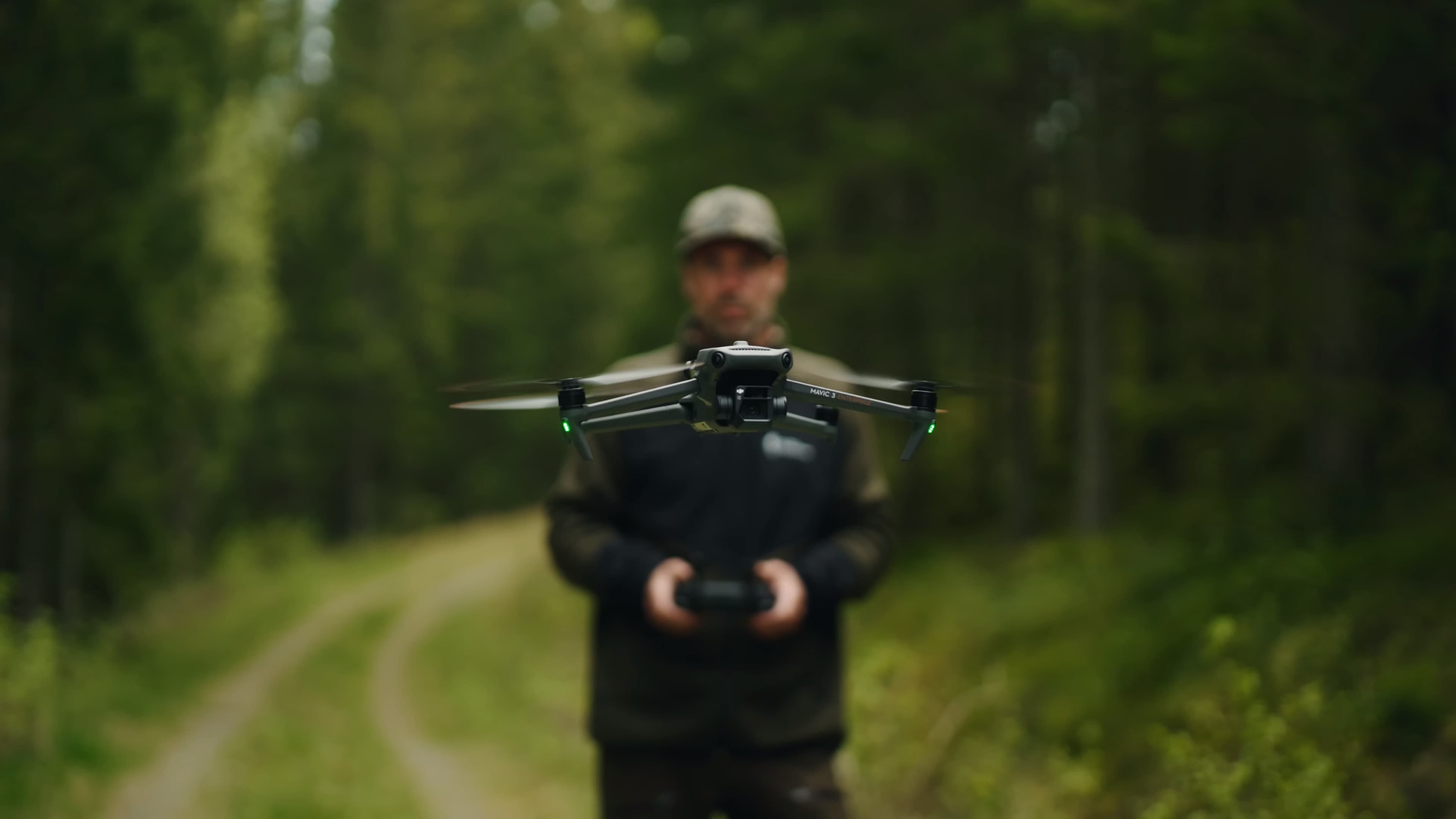 En drönare svävar framför en person som håller en fjärrkontroll, omgiven av en skogsmiljö.