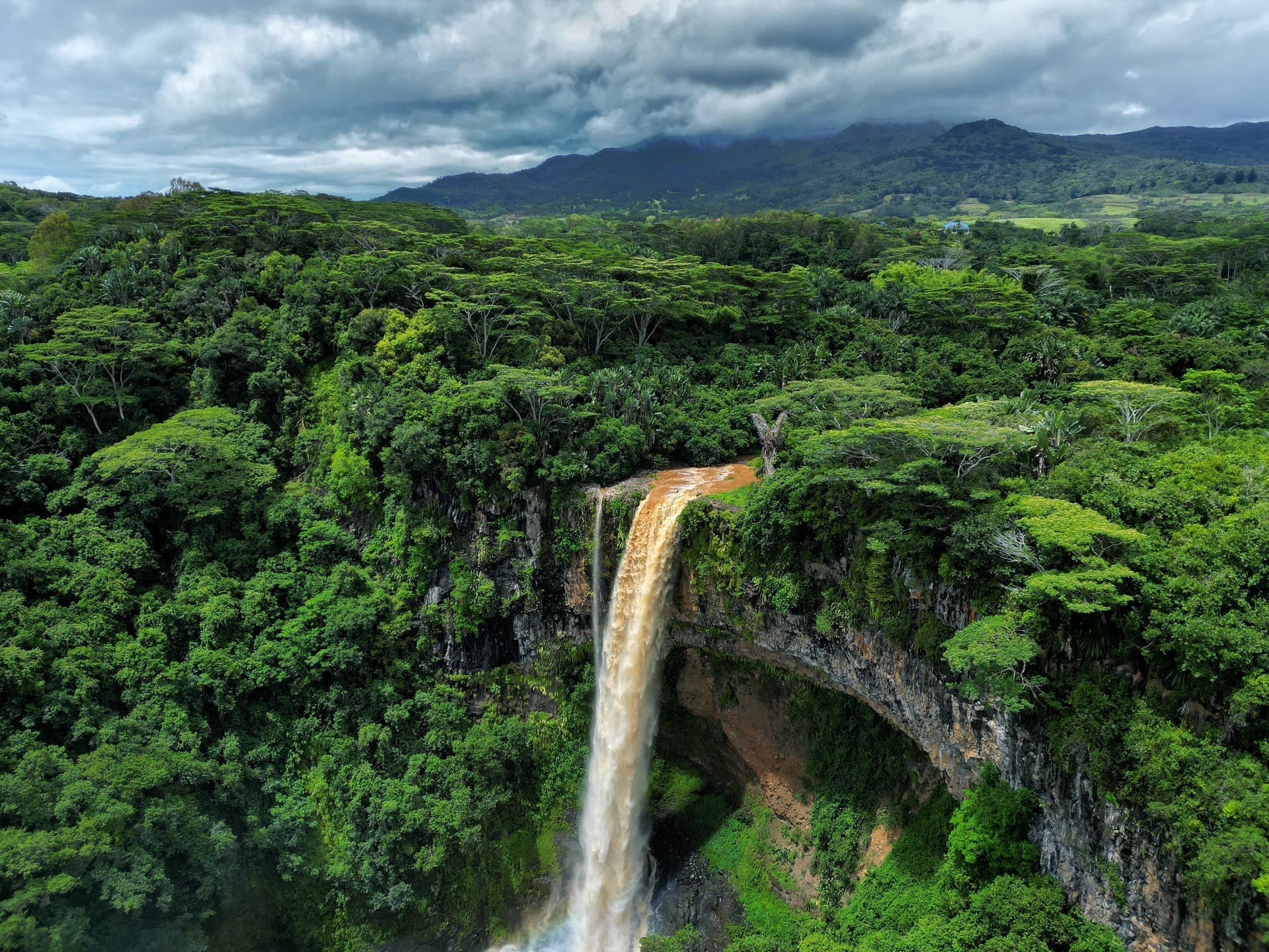 Ett majestätiskt vattenfall forsar ner från en klippa omgiven av tät, grönskande regnskog under en molnig himmel.