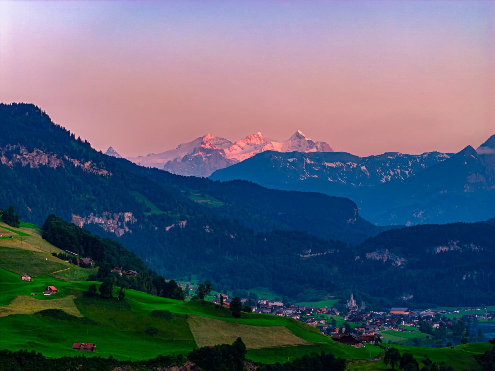 Drönarfoto taget med DJI Mavic 4 Pro: En pittoresk dal med gröna ängar och små hus, omgiven av skogsklädda berg och snötäckta toppar i solnedgångens sken.