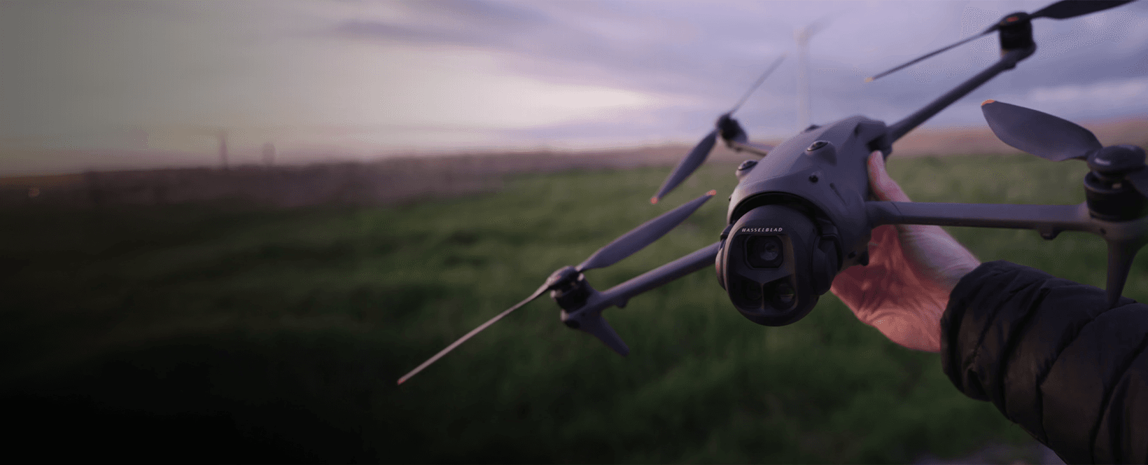 En person håller en drönare med en kamera i ett öppet landskap vid solnedgången.