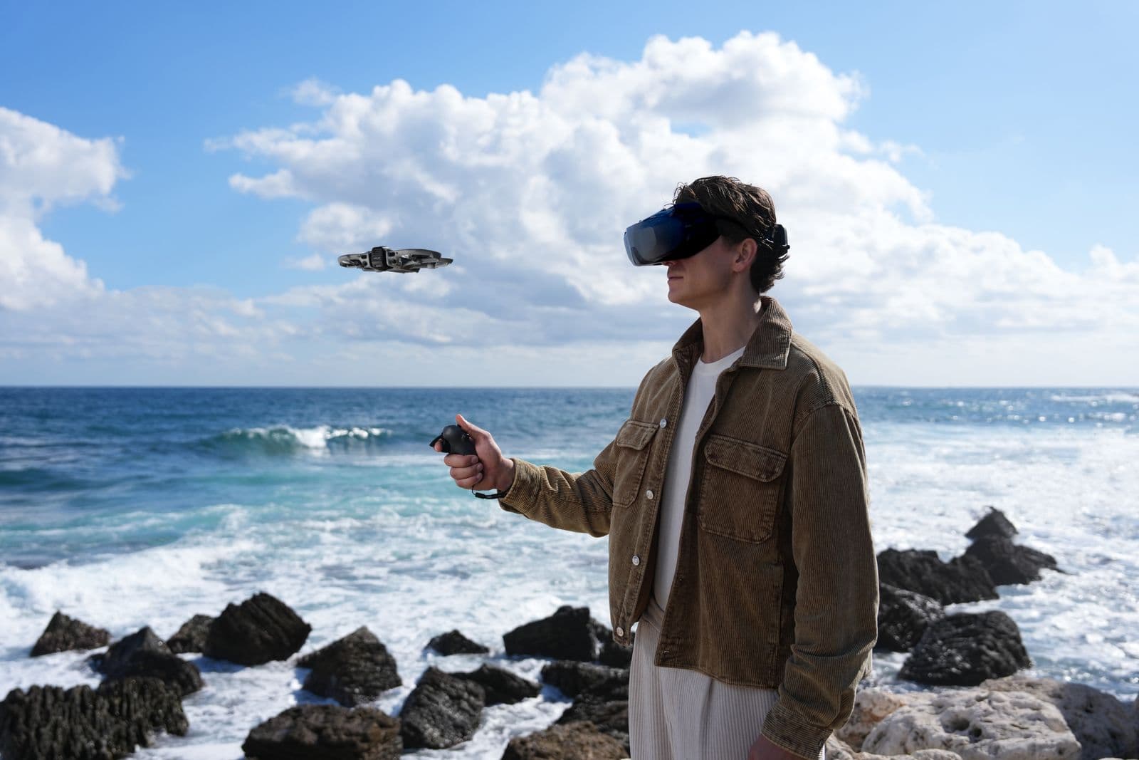 En person med VR-headset styr en DJI Avata 360-drönare vid en klippig kustlinje med havet i bakgrunden.
