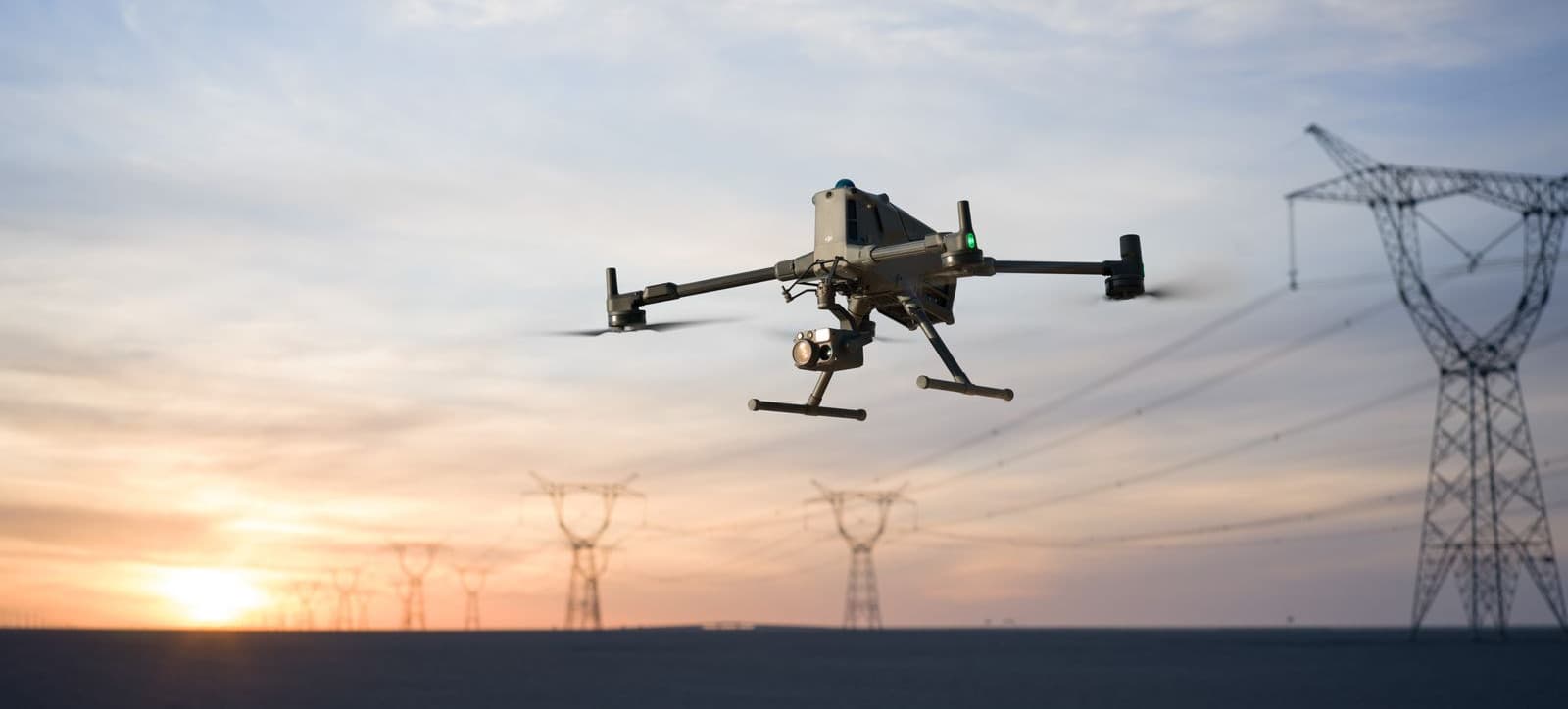 En DJI Matrice 400 svävar nära kraftledningar i solnedgången, vilket skapar en futuristisk känsla mot den färgglada himlen.