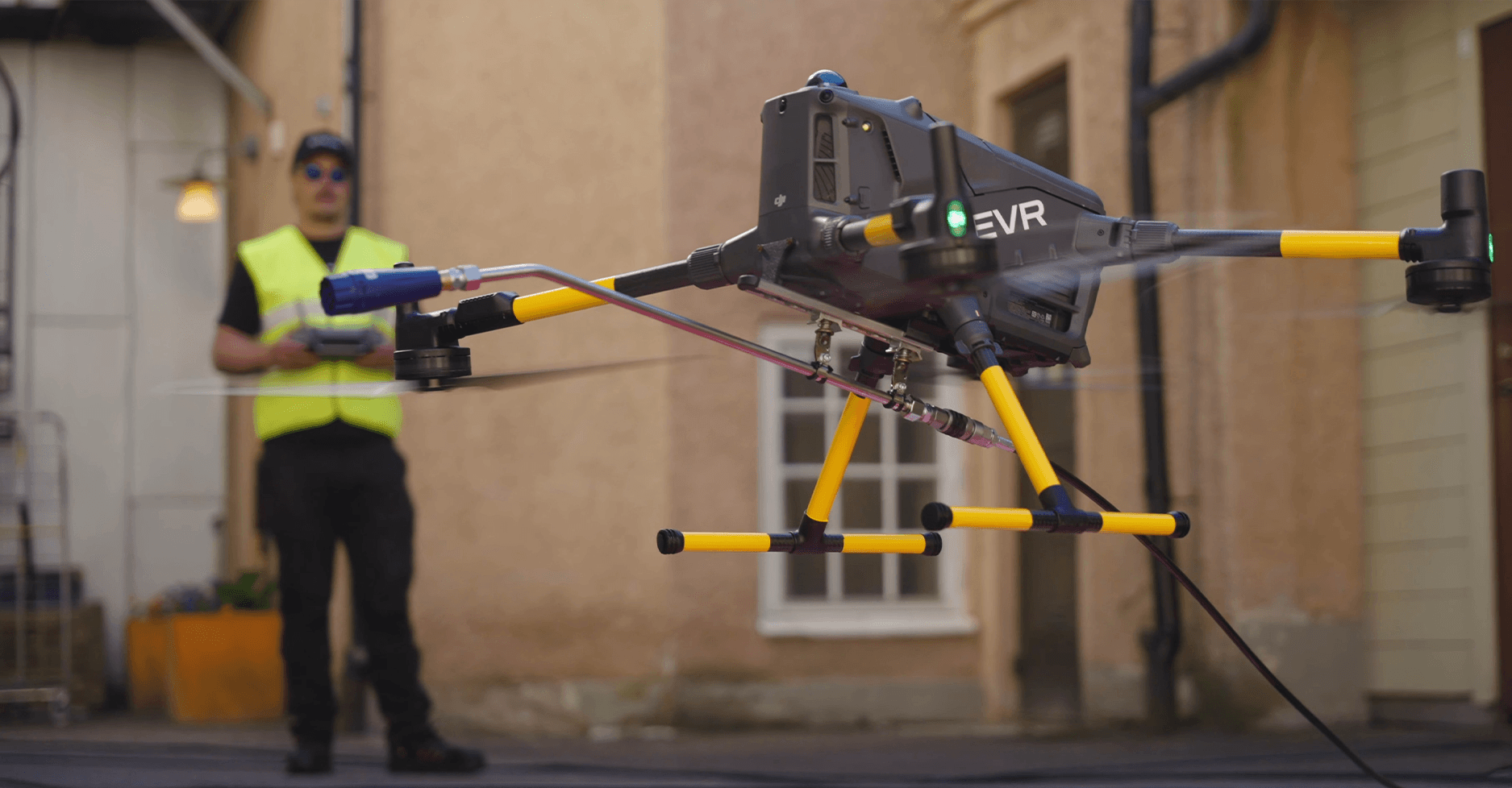 En drönare med avancerad utrustning svävar i luften medan en operatör i reflexväst styr den i en urban miljö.
