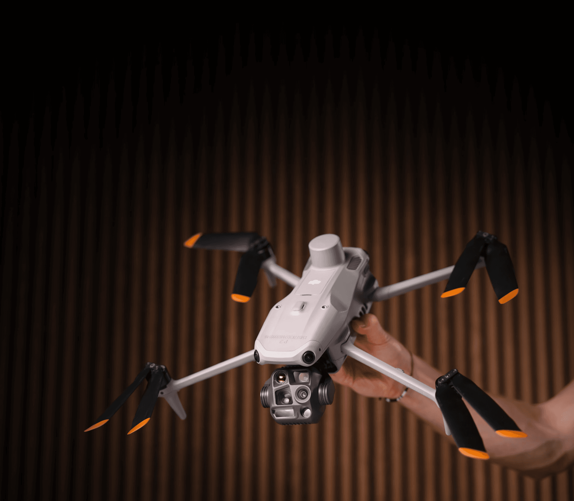 En DJI-drönare med värmekamera, en upplyst Matrice 4T hålls upp i luften av en hand med en träfärgad panel i bakgrunden.
