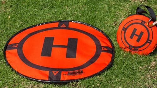 En orange Hoodman landningsplatta för drönare med ett stort 'H' ligger på gräset, bredvid en hopfällbar version i en bärväska.
