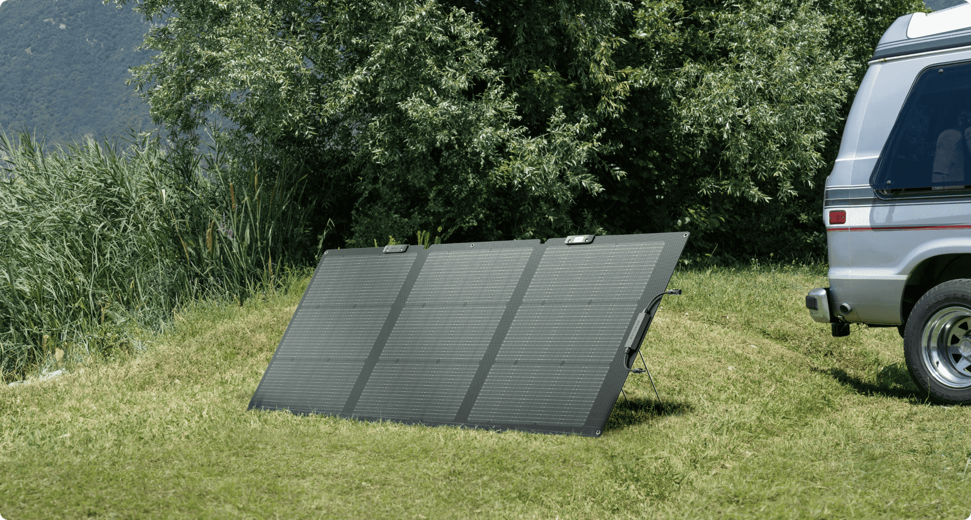 En portabel solpanel står uppställd på gräs bredvid en husbil, omgiven av grönska och träd.