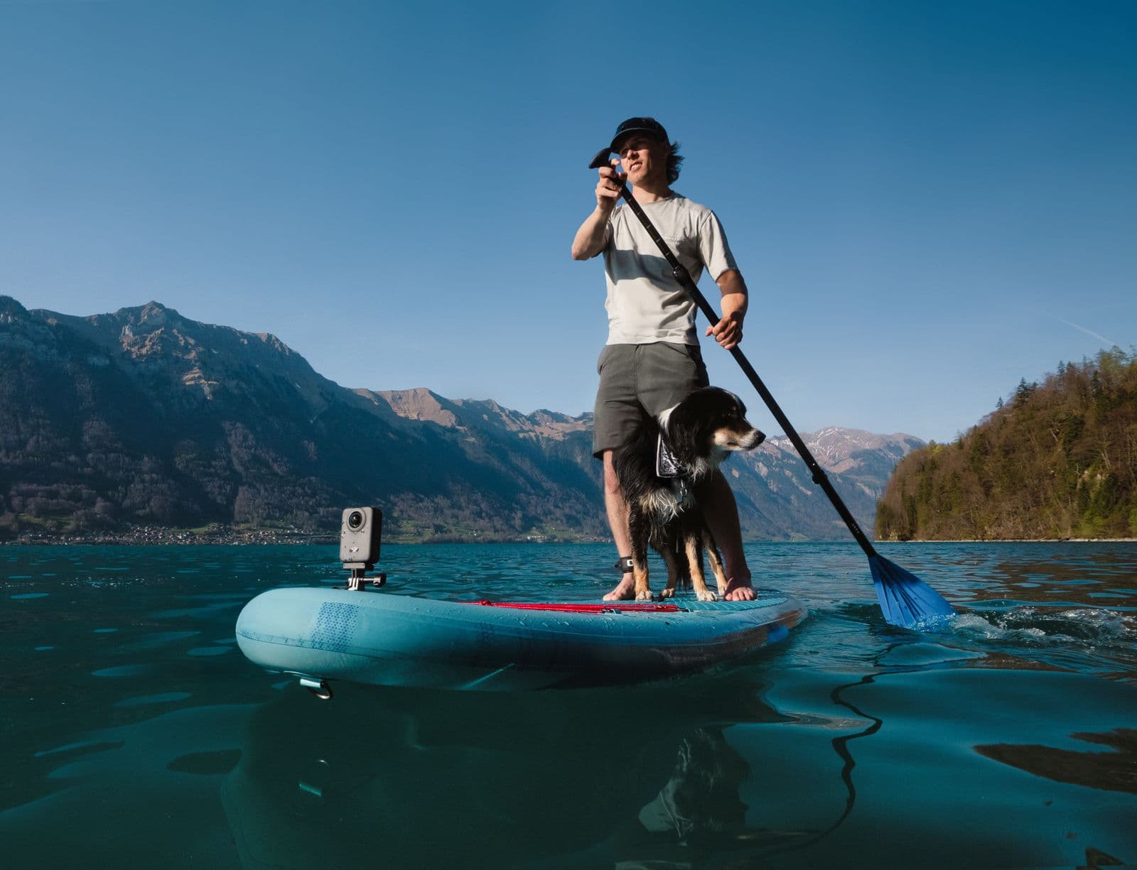 En person paddlar på en sjö med en hund på en paddleboard, omgiven av berg och klarblå himmel.