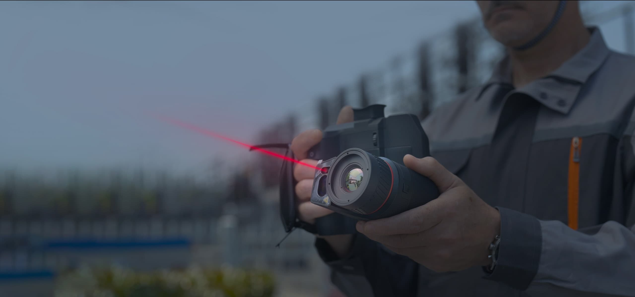 En person håller en avancerad kamera med en röd laserpekare, mot en suddig bakgrund av ett industriområde.