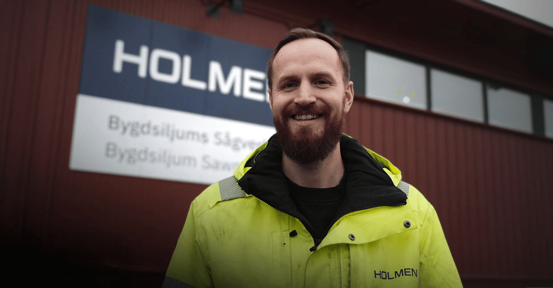 En person i en gul arbetsjacka står framför en byggnad med en skylt som säger 'Holmen'.