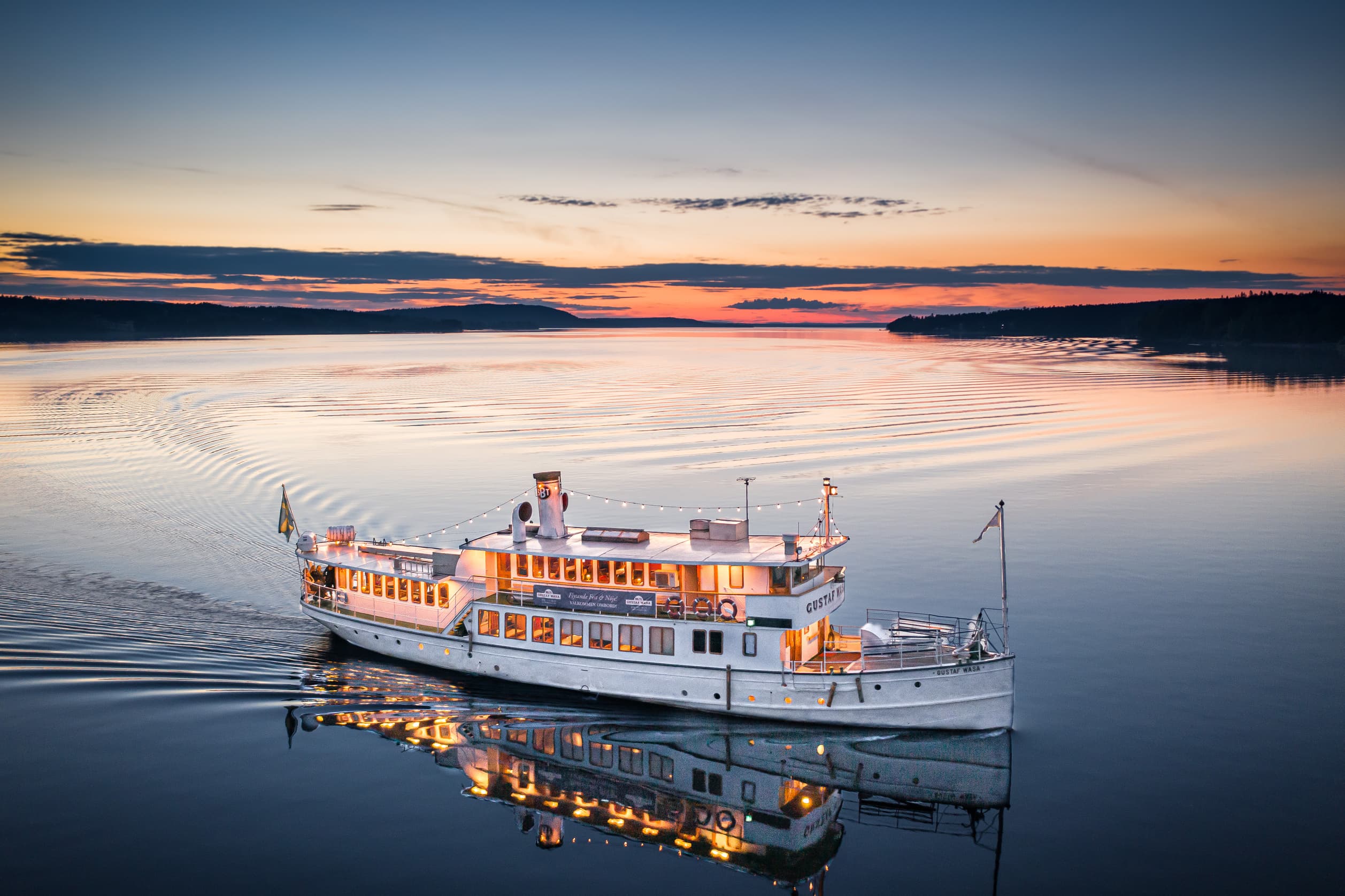Ett upplyst fartyg glider genom stilla vatten vid solnedgången, med en färgglad himmel i bakgrunden.
