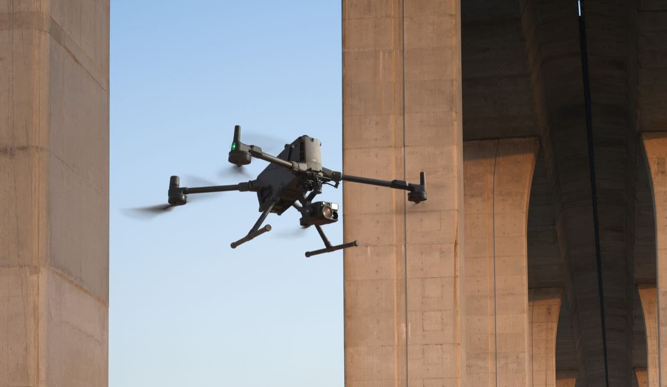 En drönare svävar nära en massiv betongbro, utförande inspektioner i en urban miljö.