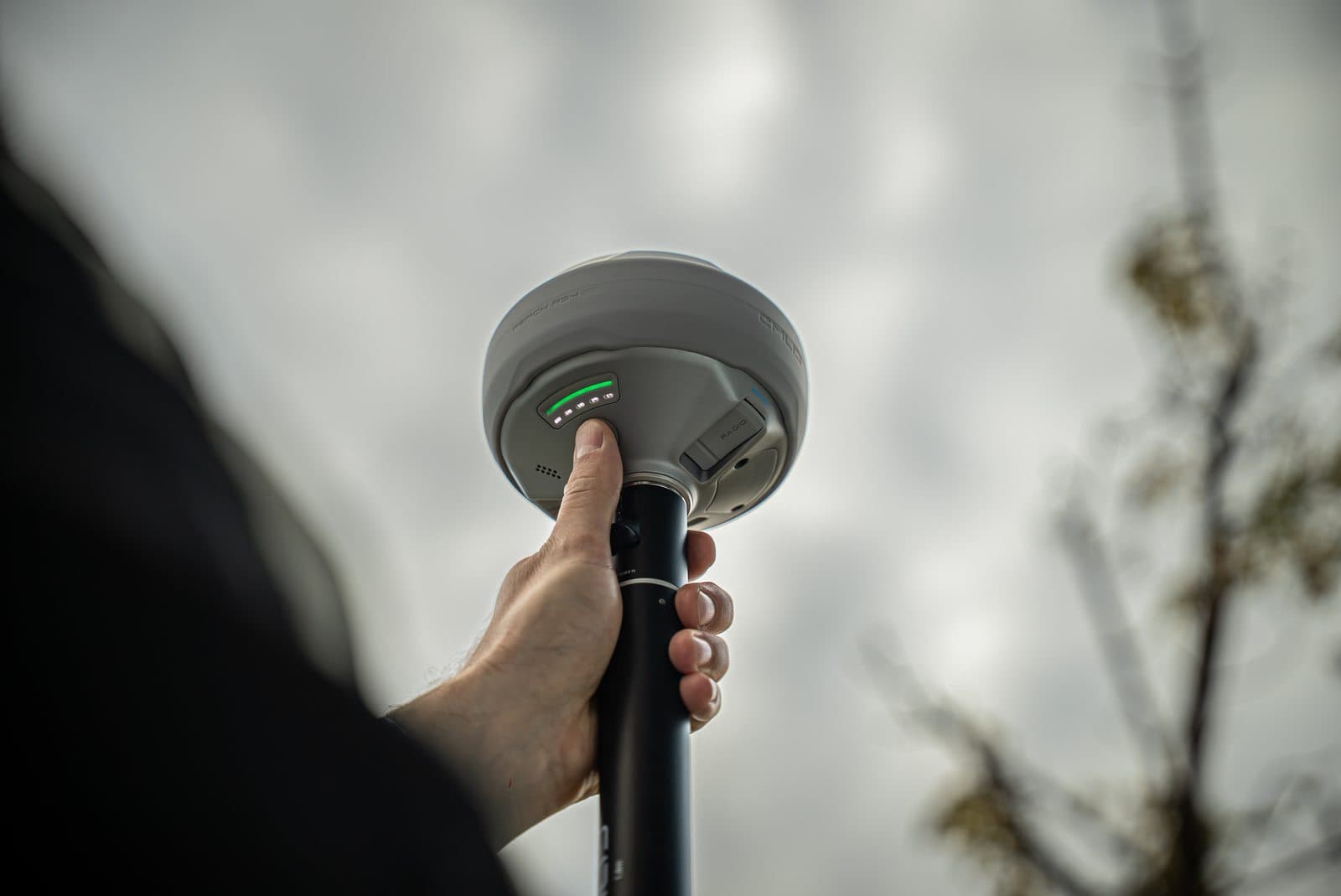 En hand håller i en Emlid Reach RS4 mot en molnig himmel, med indikatorlampor som lyser grönt.