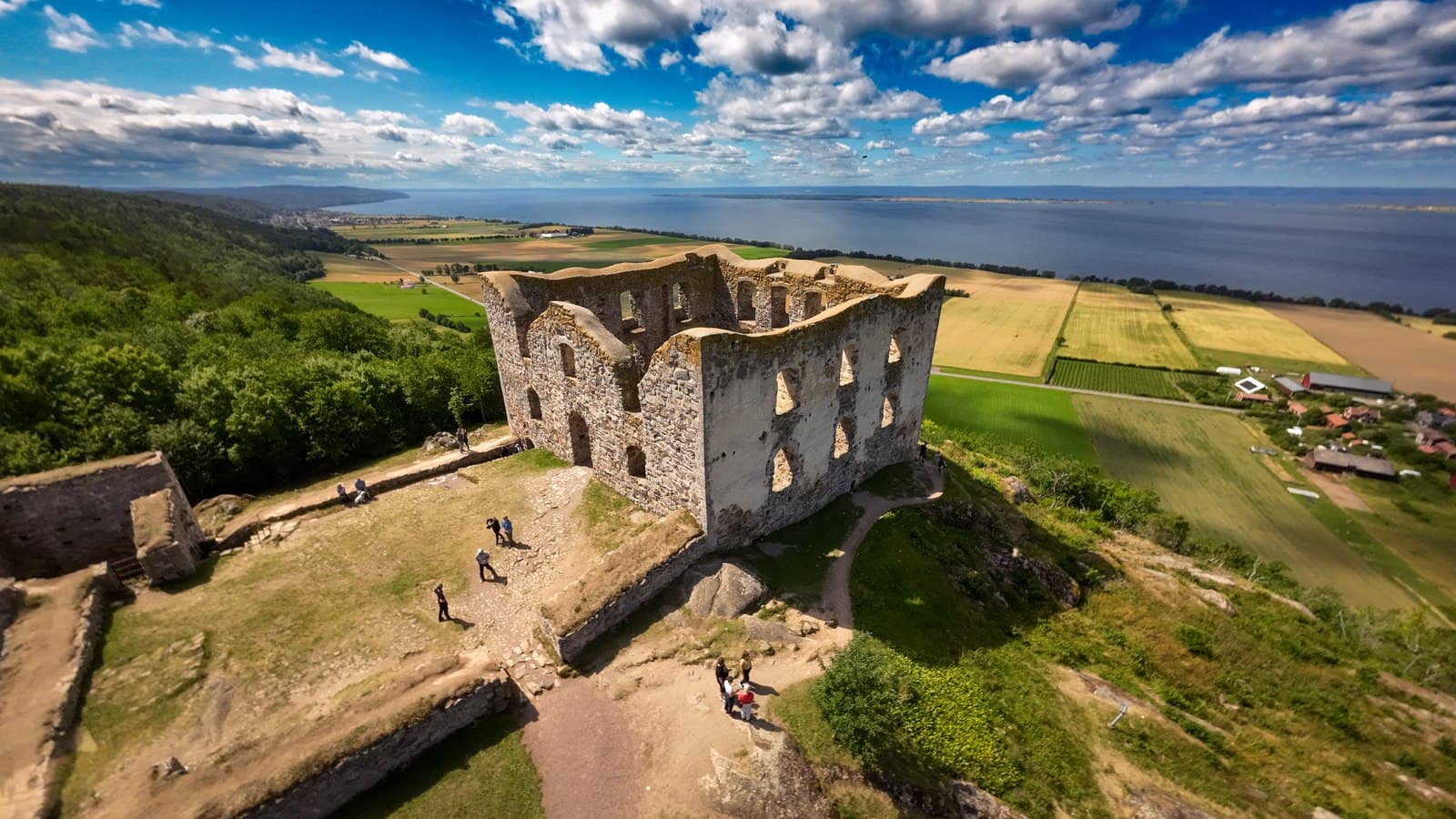 En gammal slottsruin står majestätiskt på en kulle med utsikt över ett vidsträckt landskap och en sjö i bakgrunden.