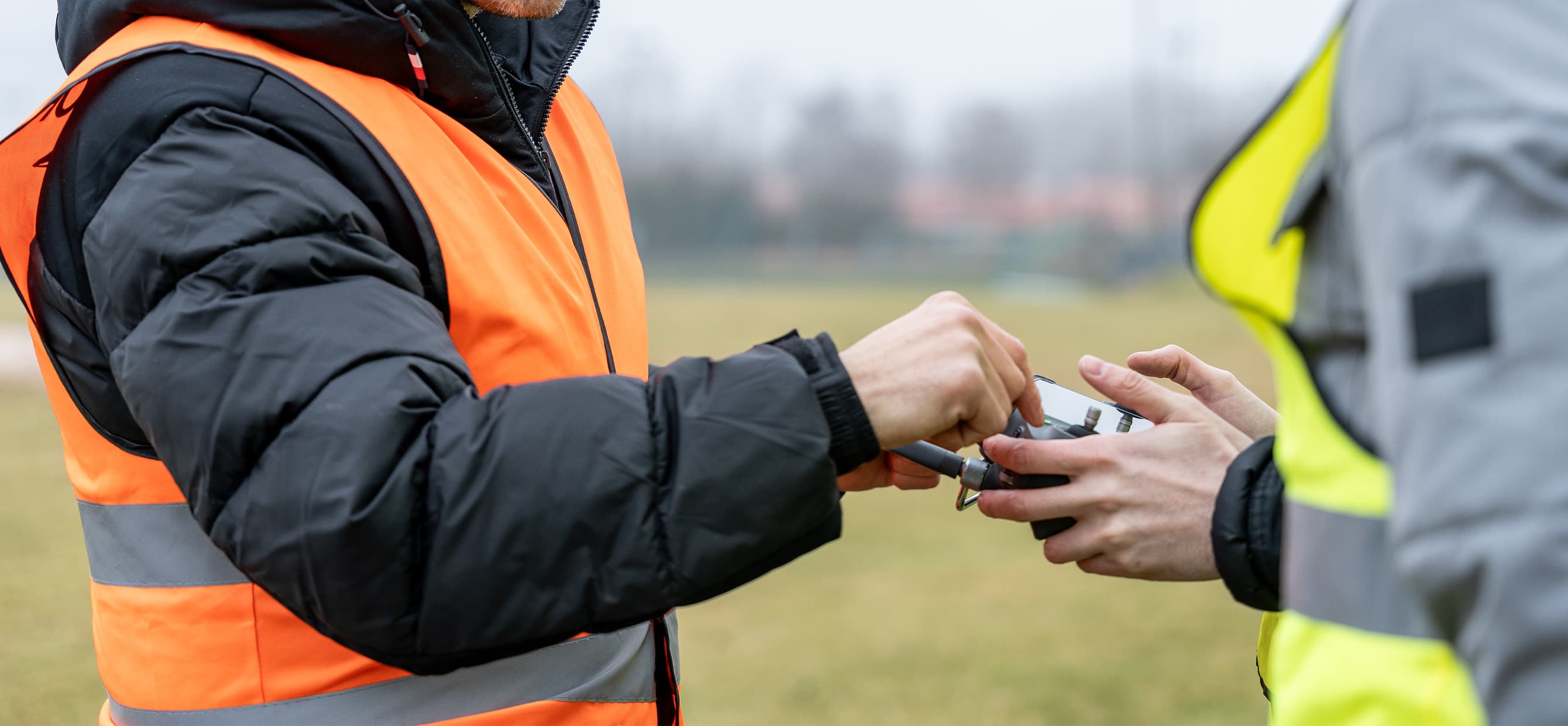Två personer i varselvästar hanterar en fjärrkontroll utomhus på en gräsbevuxen plats.