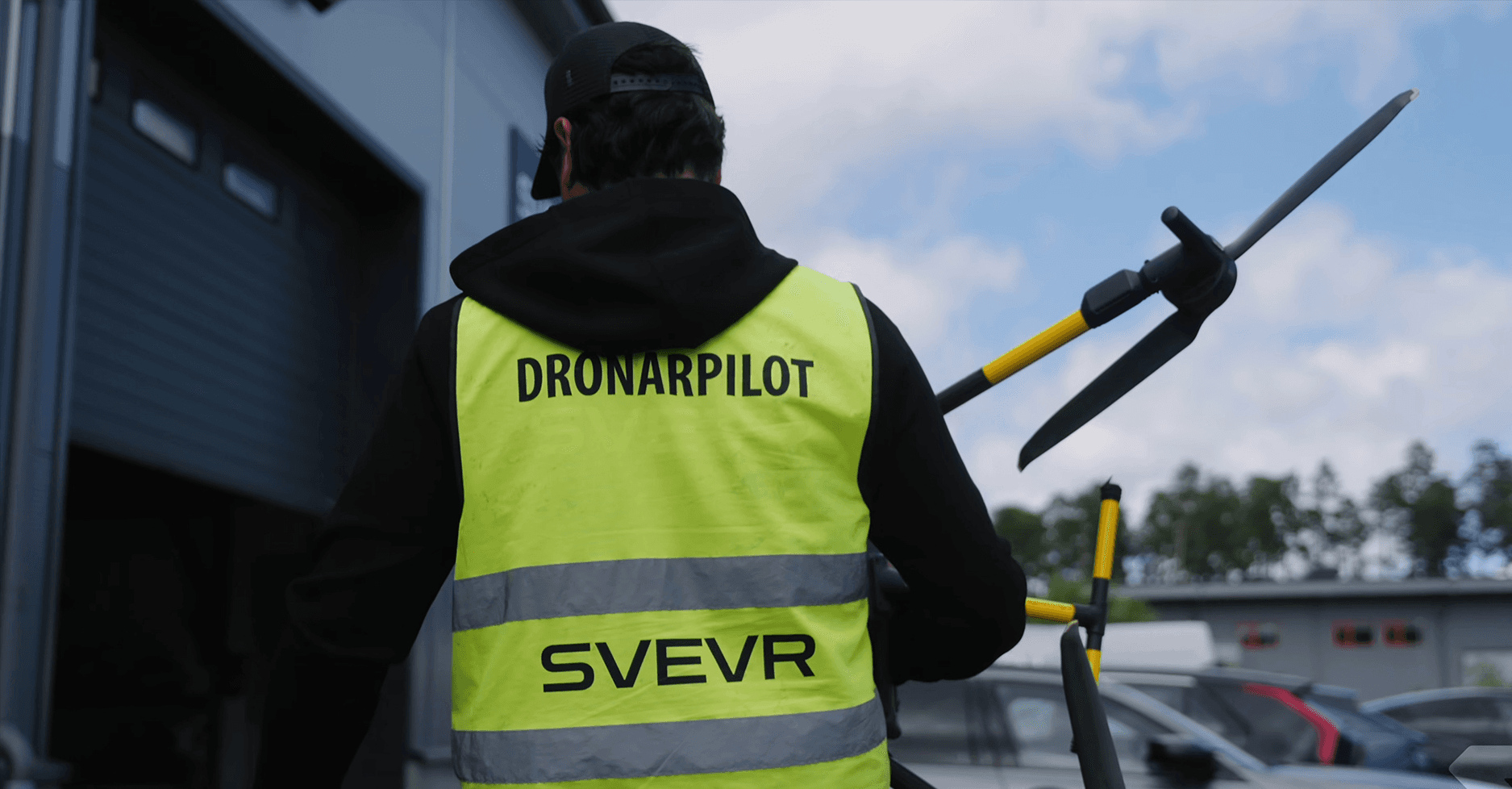 En drönarpilot i en gul väst bär en drönare utomhus, med en byggnad och bilar i bakgrunden.