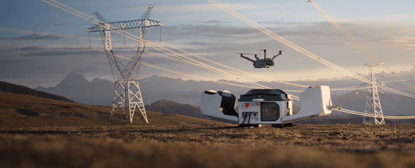 En drönare svävar nära kraftledningar i ett öppet landskap, med en laddningsstation i förgrunden.