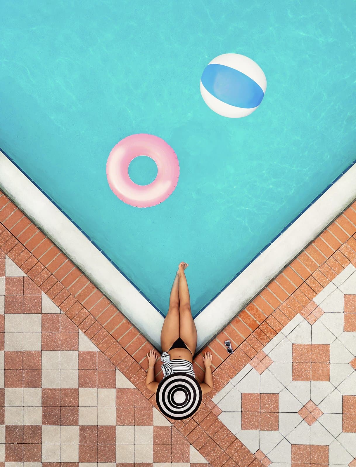 En person med en randig solhatt ligger vid kanten av en pool, omgiven av en rosa badring och en blåvit badboll.