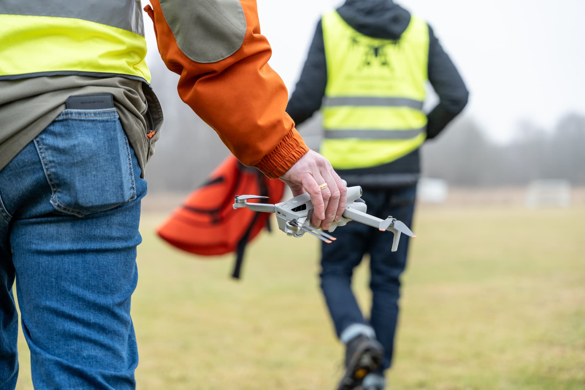 En person håller en drönare medan de går över en gräsyta, iklädda varselkläder för säkerhet.
