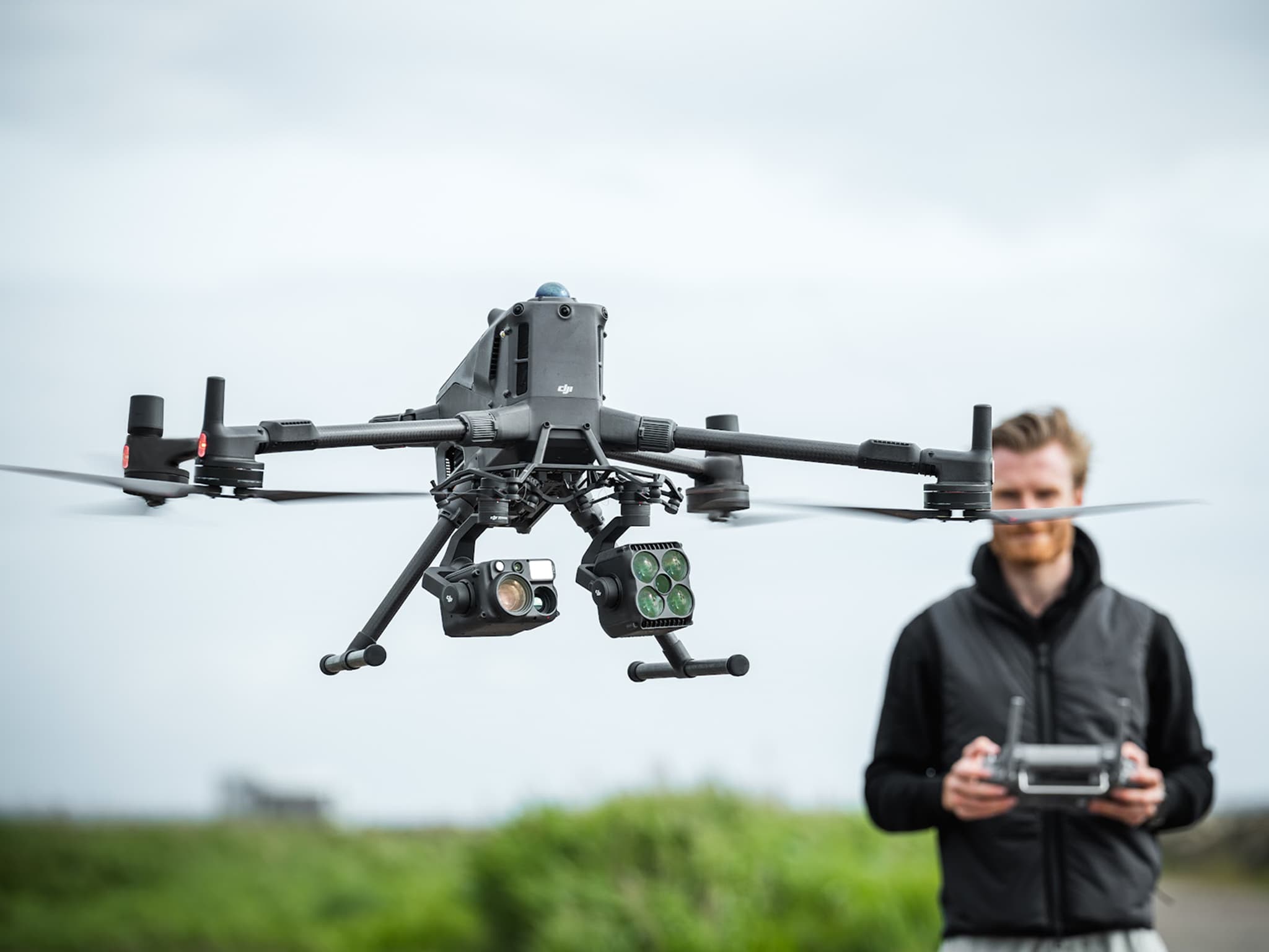 En avancerad drönare svävar i luften medan en person styr den med en fjärrkontroll i en utomhusmiljö.