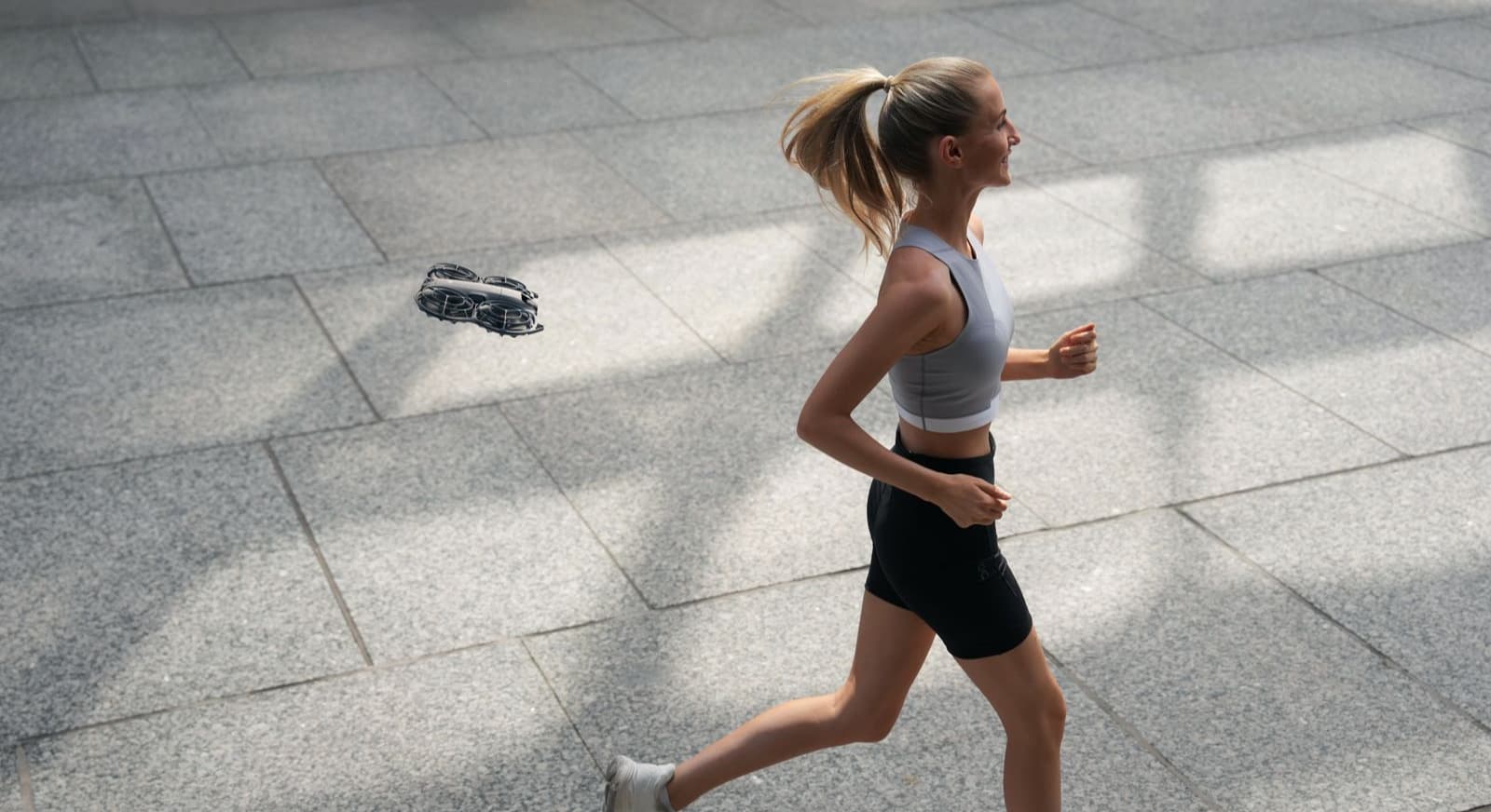 En kvinna joggar utomhus medan en liten DJI Neo 2-drönare svävar bredvid henne på en solbelyst trottoar.