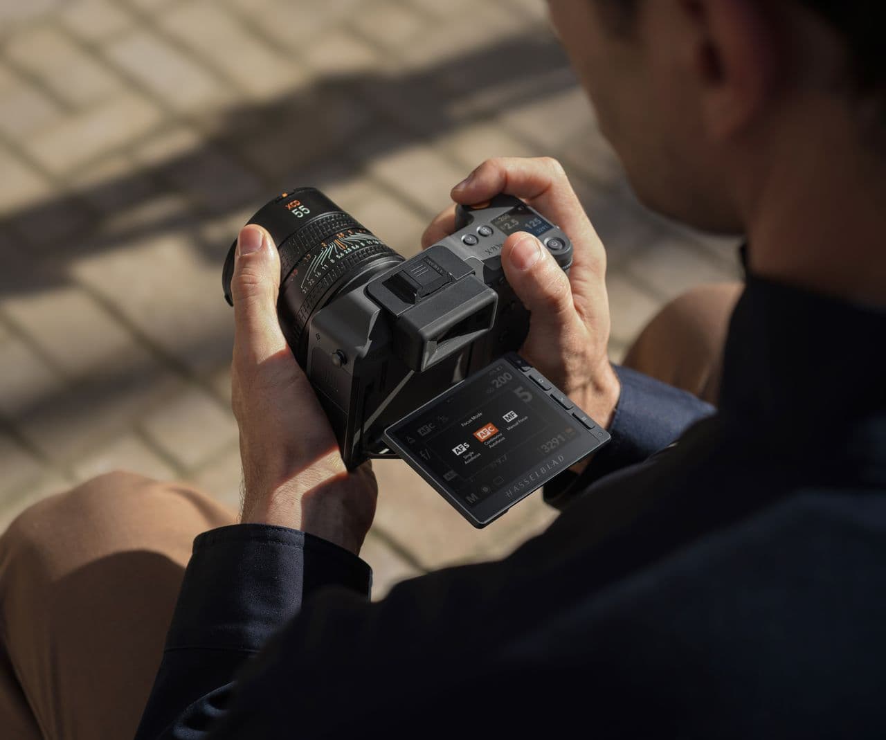 En person håller i en Hasselblad X2D II 100C kamera med en utfälld skärm, redo att ta bilder i en solbelyst utomhusmiljö.