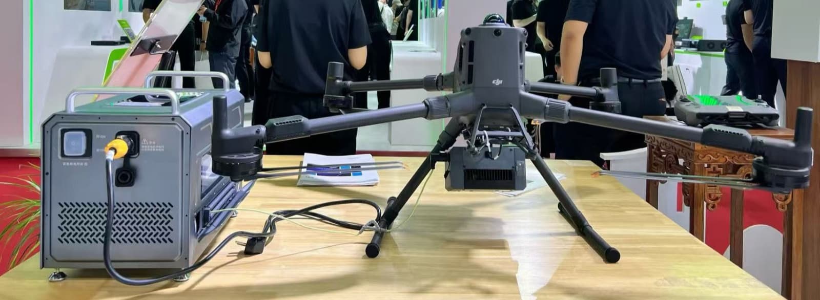 En avancerad drönare visas upp på ett bord vid en teknikutställning, omgiven av besökare och teknisk utrustning.