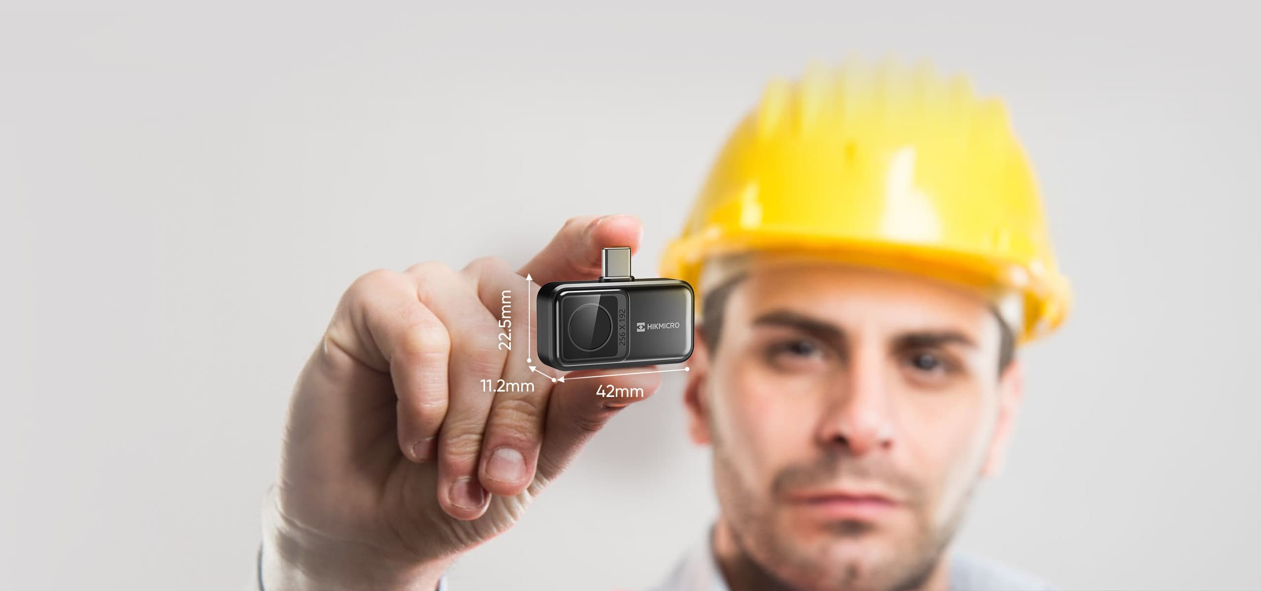 En person med gul hjälm håller upp en liten teknisk enhet med angivna mått, mot en neutral bakgrund.