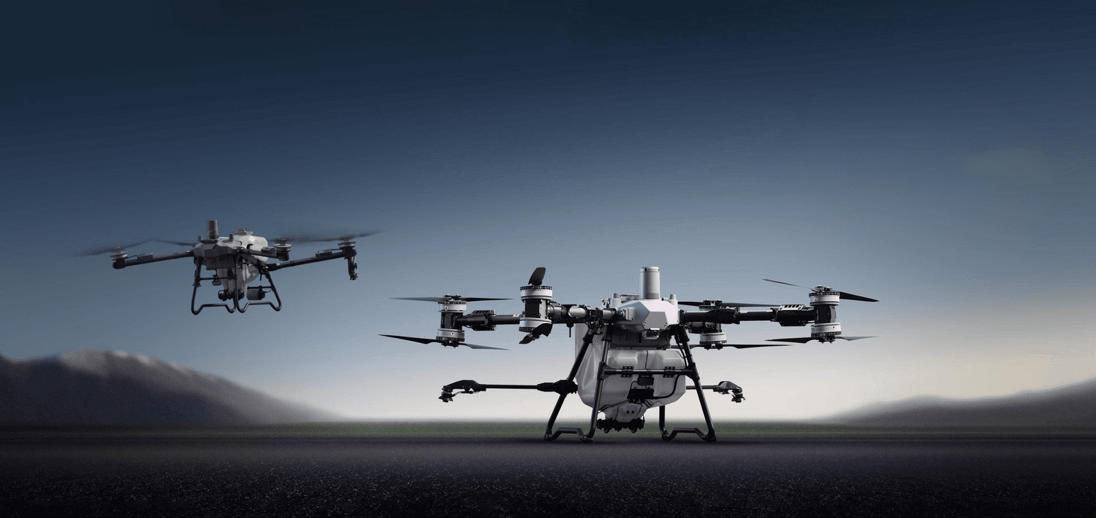 Två drönare svävar över en öppen yta med en bergig horisont i bakgrunden, redo för uppdrag i skymningen. T70P till vänster, T100 till höger