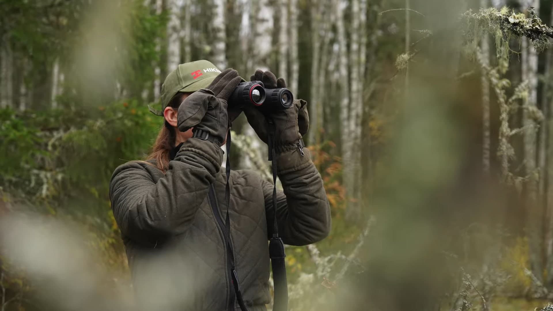 En jägare i grön jacka och keps spanar med en värmekikare i en höstig björkskog.