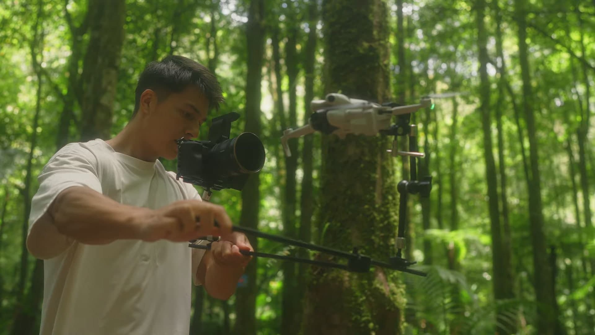 En filmare i regnskog håller en kamera och en lång POV-arm med en liten DJI-drönare fäst i änden.