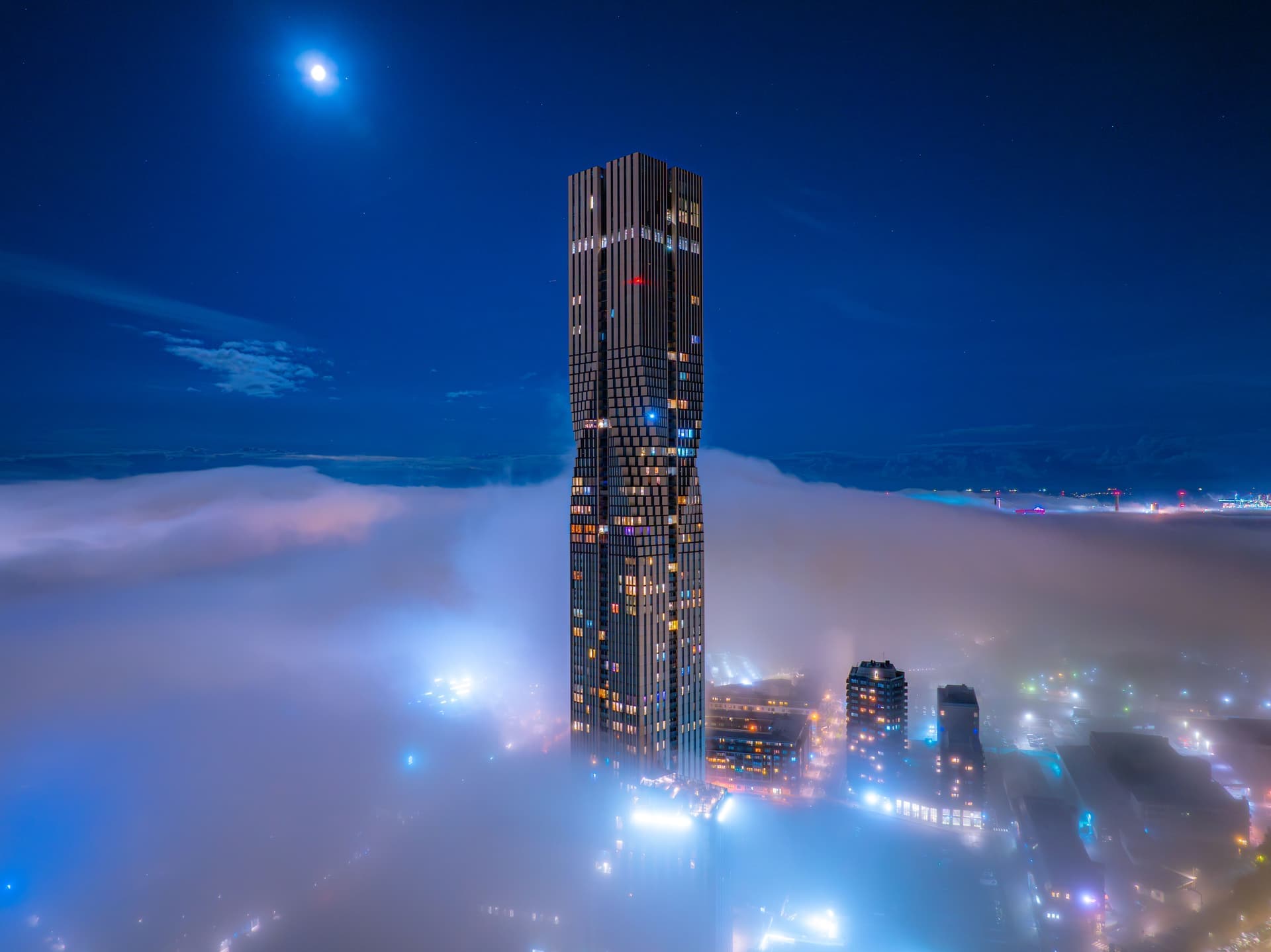 En modern skyskrapa reser sig genom dimman i en upplyst stad under en klar natthimmel med månen synlig ovanför.
